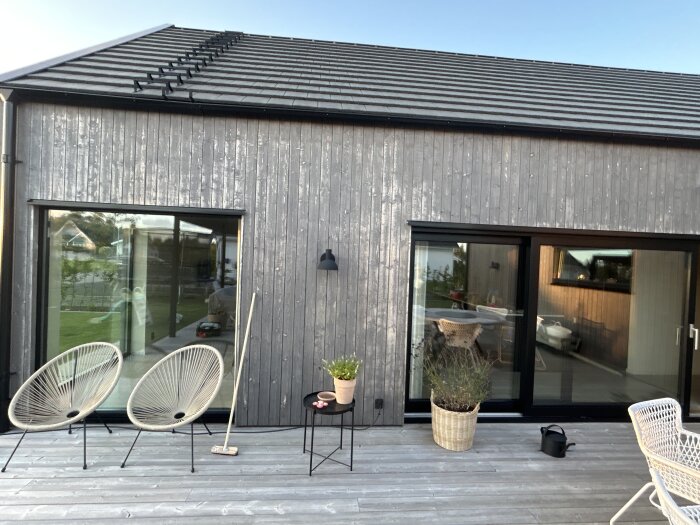 Grått enkelplanshus med två stora fönster och altan, två stolar och ett bord med växt. Panelen ser väderpåverkad ut.