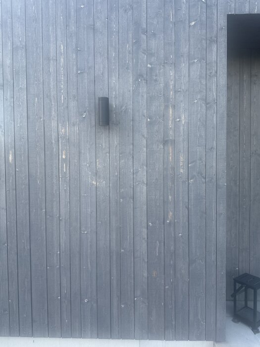 Grå träfasad med lasyrproblem, exempelvis fläckar, syns på en husvägg med ett svart väggljus och en lanternin längst ned i hörnet.