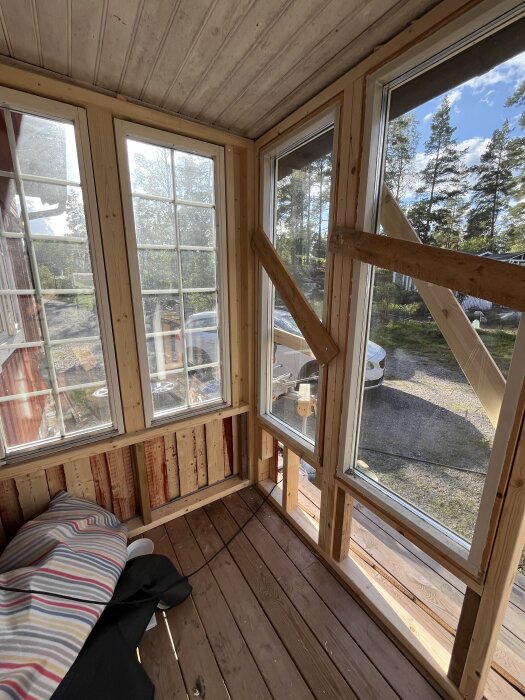 Träveranda med stora glasfönster i ram, sedd från insidan. Fönstren har spröjsade rutor och omgivningen utanför visar träd och himmel.
