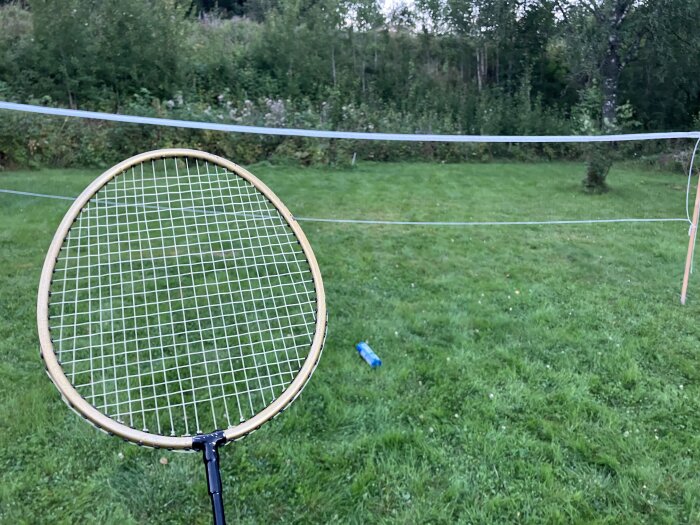Badmintonracket och nät på en gräsmatta i en trädgård, omgiven av träd.