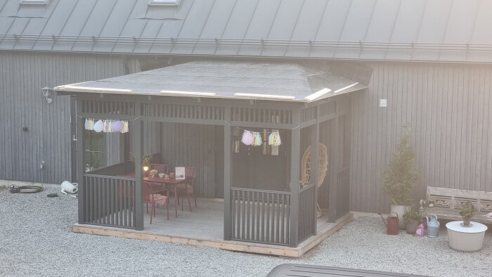 En veranda med ett nylagt pappklätt tak och enkel inredning, sedd utifrån, omgiven av några trädgårdsredskap och möbler på en grusad yta.