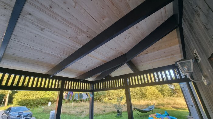 Slätspontat tak under konstruktion, med snedställda svarta takstolar och en vy över trädgård med lekredskap och bil i bakgrunden.