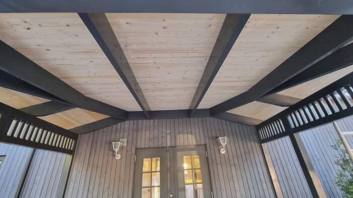 Tak av slätspont och takstolar, med delvis synliga vägglampor och dörrar. Bilden tagen i motljus med en suddig effekt.