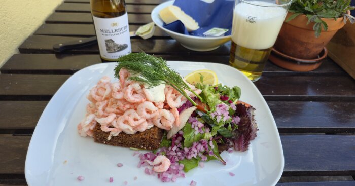 Räksmörgås med dill, citron, rödlök och sallad på en tallrik, serverad med en alkoholfri ölflaska på träbord.