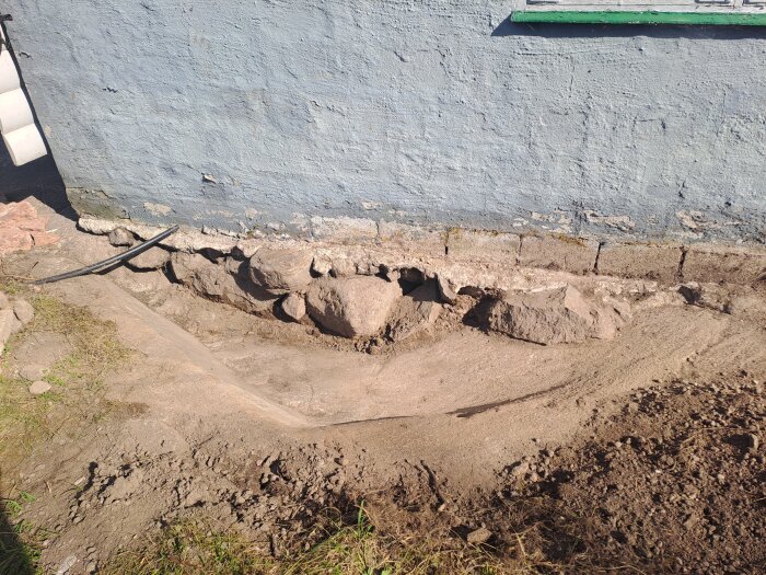 Grop med större stenar intill en husgrund, del av åtgärd för fuktskador på suterräng-källare, förberedd för grovbetong och dränering.