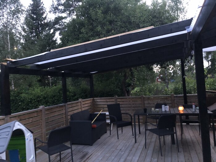 Altantak med segelduk över en uteplats, omgiven av träd och staket, med utemöbler och ett tänd ljus på bordet.