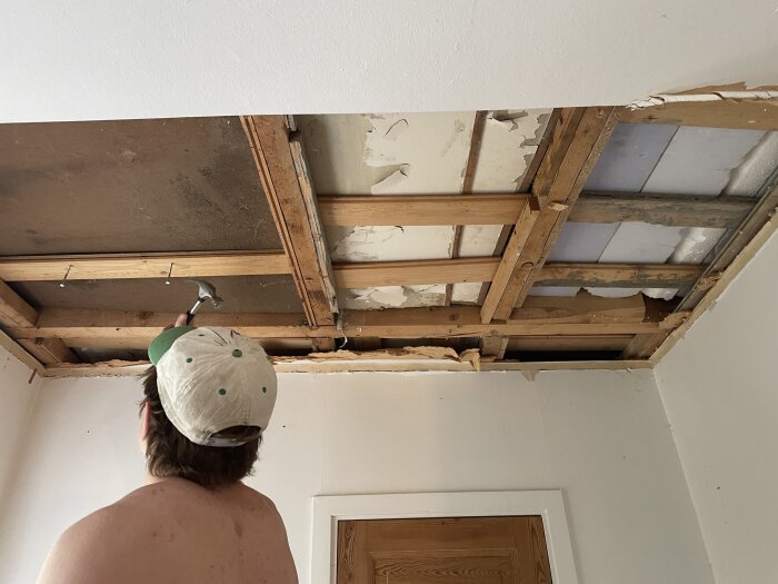 Person utan tröja med keps arbetar med en hammare på ett tak under renovering i ett gammalt hus.