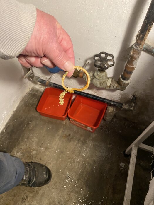 En hand håller ett avbrutet snöre framför en gammal vattenkran med rör och röda hinkar på golvet.