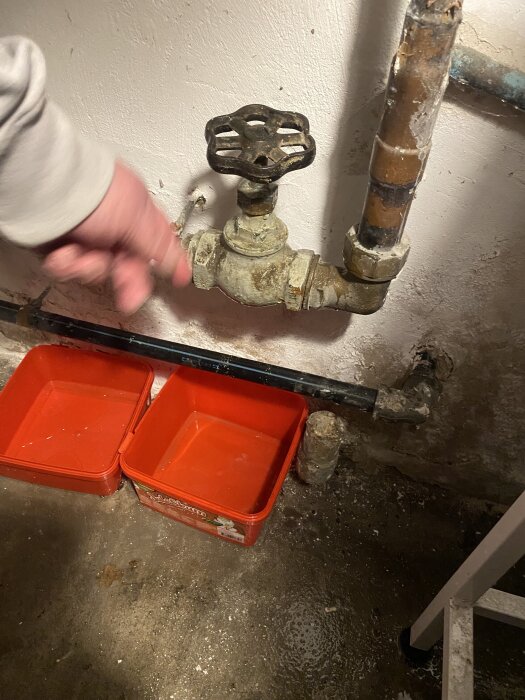 Äldre vattenkran med rostiga rör och en hand som pekar, två röda plastbehållare på golvet under för att samla upp eventuellt läckage.