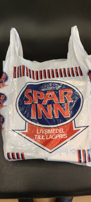 Plastpåse med texten "Spar Inn" och "Livsmedel till lågpris", vit påse med röda och blå ränder på handtagen, liggande på svart yta.
