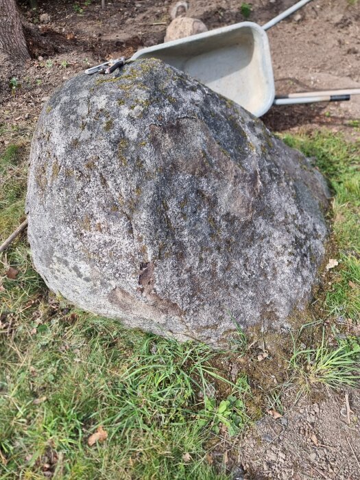 Stor sten på gräsmatta med en skottkärra i bakgrunden, verktyg placerade ovanpå stenen.