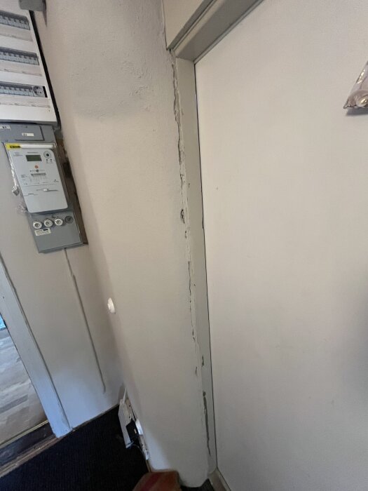 Sprukket murpuss ved siden av ytterdørkarmen. Hullet er synlig med ujevn kant og noen kabler til venstre på veggen.