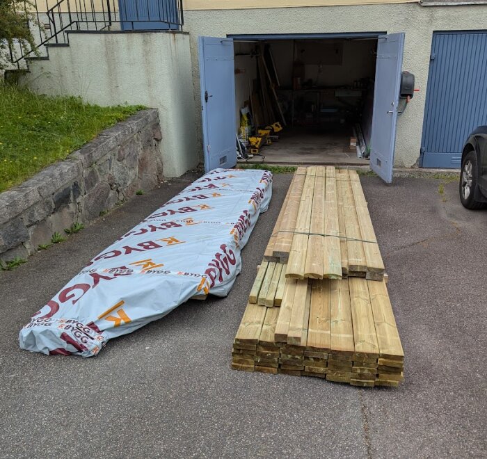 Brädor och byggmaterial levererat på en uppfart framför en garageport, redo för altanprojekt.