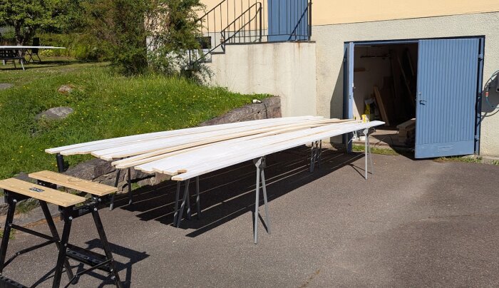 Målade träplankor torkar på bockar utomhus framför ett öppet förråd under ett altanrenoveringsprojekt.