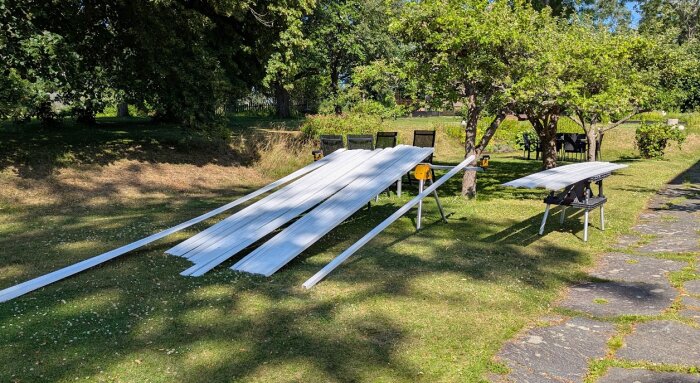 Målade träplankor placerade på bockar utomhus i en trädgård, redo för bygge av altantak och räcke, omgivna av gräs och träd i bakgrunden.