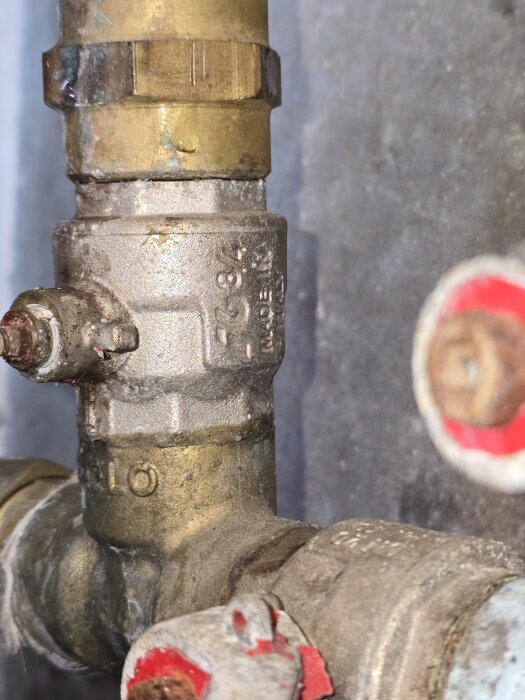 En närbild av en äldre kulventil i metall som är fastmonterad i ett rörsystem, med skadade och avskavda delar.
