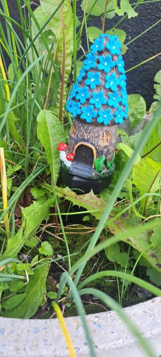 Minatyrhus med blåa blommor på taket och röda svampar vid sidan, placerat bland grönska i en trädgårdsmiljö.