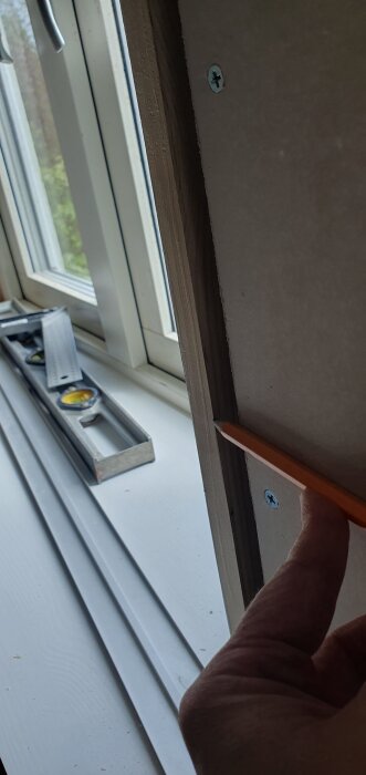 En fönstersmyg, där en hand håller en penna mot kanten för att mäta millimeterskillnad mot gipsväggen. Ett vattenpass syns i bakgrunden.