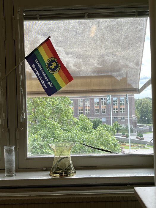 Regnbågsflagga i fönster, med texten "Idrottsrörelsen gör Sverige starkare". Utsikt över träd och en byggnad i bakgrunden.