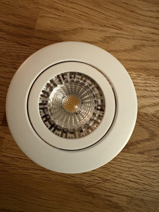 Vit spotlight med rund ram, placerad på ett trägolv. Omgiven av diskussion om byte av trasiga badrumsbelysningar.