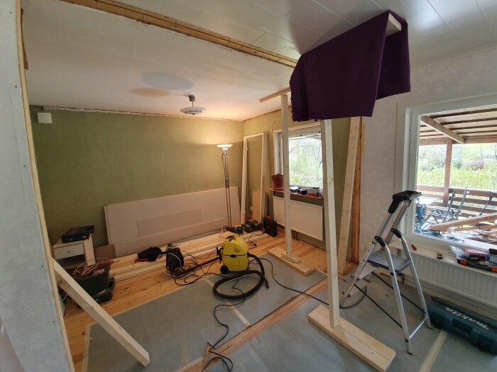 Rum under renovering med stödreglar och verktyg, dammsugare och stege inomhus bredvid ett fönster, för att förhindra att golvet sätter sig.