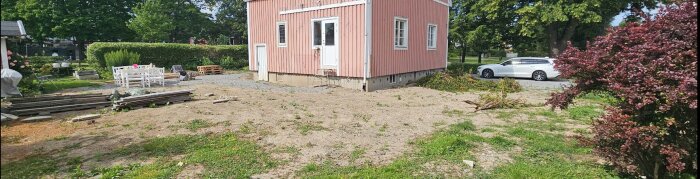 Gräsmatta framför ett rosa hus, delvis avgrävd efter dränering. Trädgårdsmöbler och material på marken, bil parkerad i bakgrunden.