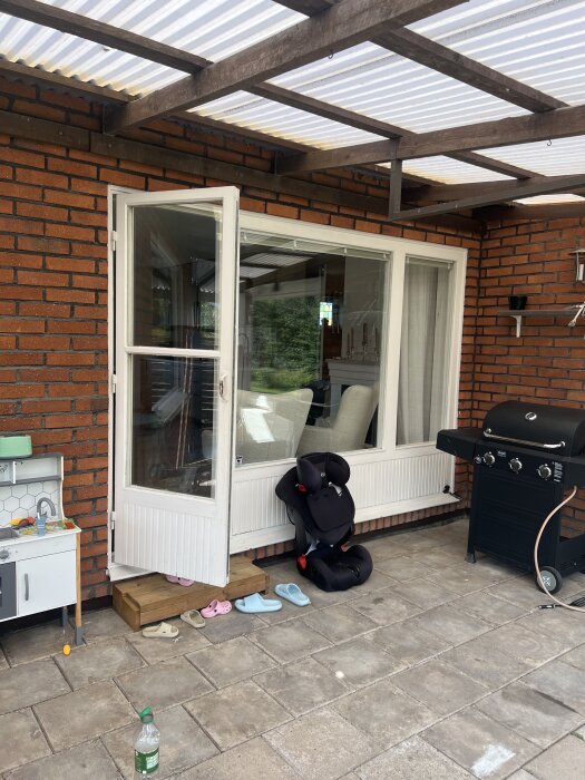 Tegelhus med altan, vit dörr och fönster, barnstol, grill och skor på marken.