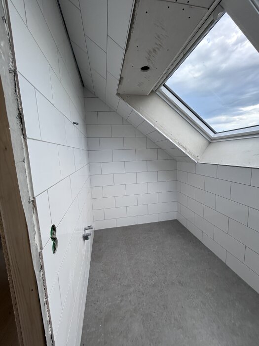 Färdigkaklat badrum med vitkaklade väggar, grått golv och takfönster som släpper in naturligt ljus.