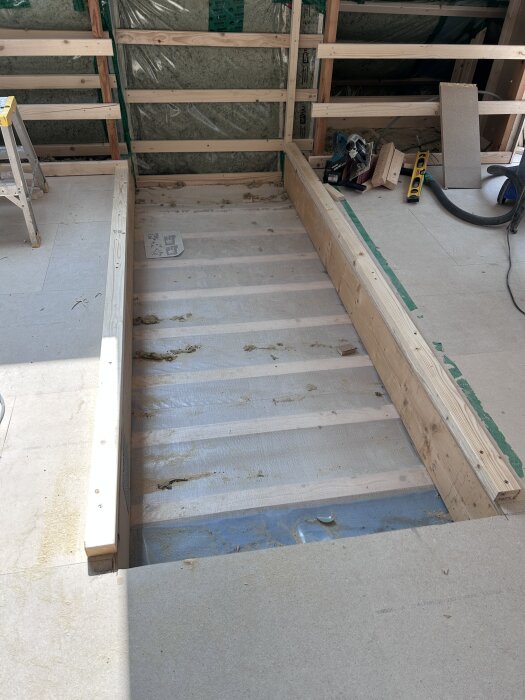 Öppning i golvet med träreglar för trappa under byggnation i ett hus. Bakgrund visar isolering och verktyg.