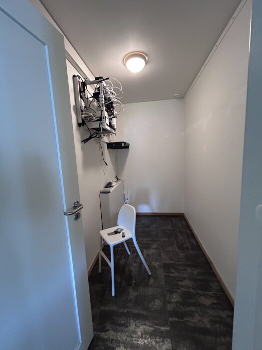 Tomt rum med stängd vit dörr, svart golv, en stol och teknikprylar på väggen under en taklampa.