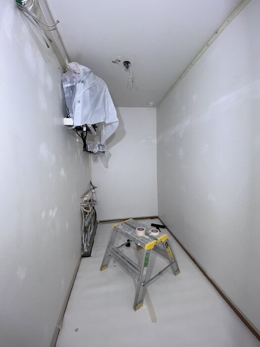 Ommålat rum under renovering, med stege och tejprullar på golvet samt synliga rör och elektriska kablar på vägg och tak.