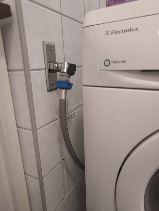 Ballofixventil med lös koppling vid tvättmaskin, slangen pekar bort från maskinens baksida. Placering på väggplatta vid vit tvättmaskin.