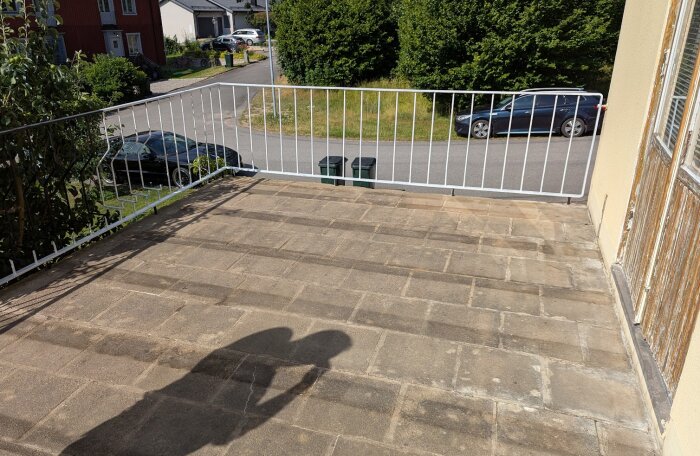 Gammal altan med stenplattor och vitt räcke. Skugga av person syns på plattorna, omgivning med gräsmatta, väg och bilar i bakgrunden.