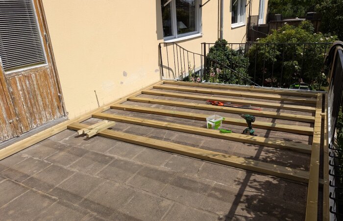 Träram för ny altan byggd på stenlagd yta, verktyg och byggmaterial synliga, del av husfasad och trädgård i bakgrunden.