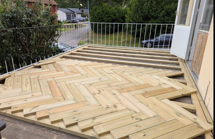 Nybyggd altan med trämönster i fiskbensläggning och delvis färdigt golv, omgiven av vitt räcke och trädgårdsutsikt.