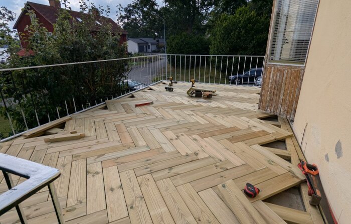Nybyggd altan i trä med fiskbensmönster som delvis är färdigställd, omgiven av ett vitt stålstaket. Verktyg ligger på altanen.