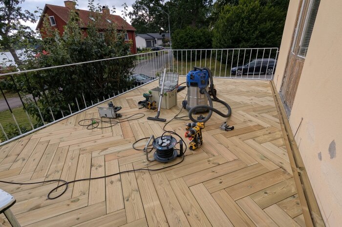 Nybyggd altan med trämönster, diverse verktyg och utrustning syns, inklusive borrmaskin och dammsugare. Hus och träd i bakgrunden.