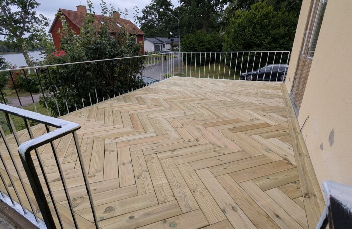 Nybyggd altan med trämönster i fiskbensläggning och metallräcke, omgiven av hus och grönska.