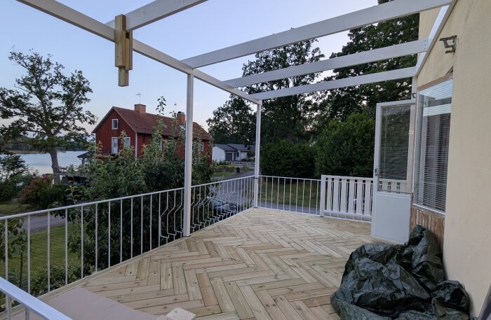 Nybyggd altan med trägolv i fiskbensmönster och vita räcken, omgiven av träd och bostäder, med utsikt över ett vattendrag och ett rött hus i bakgrunden.