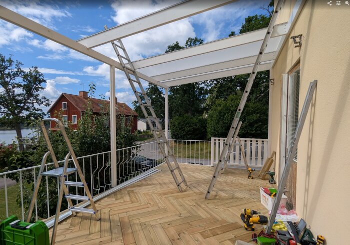 Altan under konstruktion med trätrall i fiskbensmönster, stegar lutar mot en struktur med kanalplasttak. Verktyg och grön låda syns på altanen.