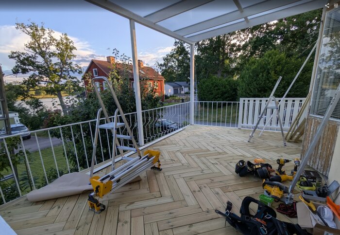 Nybyggd altan med trätrall, verktyg och stegar, omgiven av träd och hus i bakgrunden, under en solig himmel.