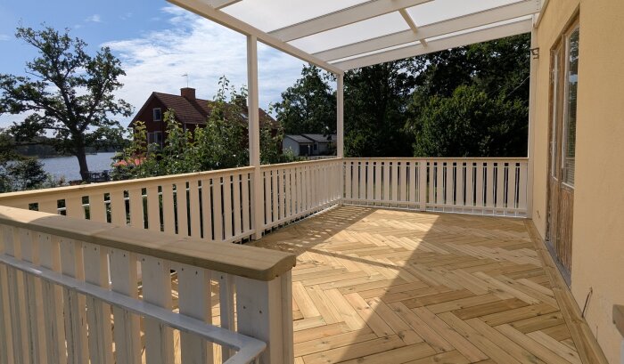 Nybyggd träaltan med vita räcken och tak av kanalplast. Utsikt över trädgård med villa, sjö och träd i bakgrunden under solig himmel.
