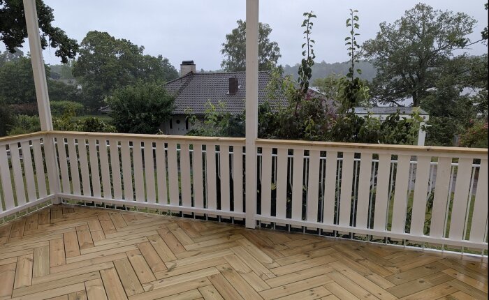 Altan med nybyggt trätrall och vitt räcke i trädgård, omgiven av grönska och träd i bakgrunden.