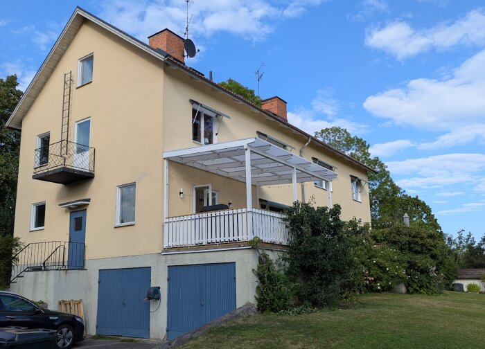 Gul villa med vitmålad altan under tak, bredvid blå garageportar och grönskande trädgård.