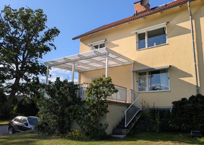 Hus med nybyggd altan i den övre våningen, vit takstruktur och trappa ner till trädgården; solig dag och grönskande omgivning.