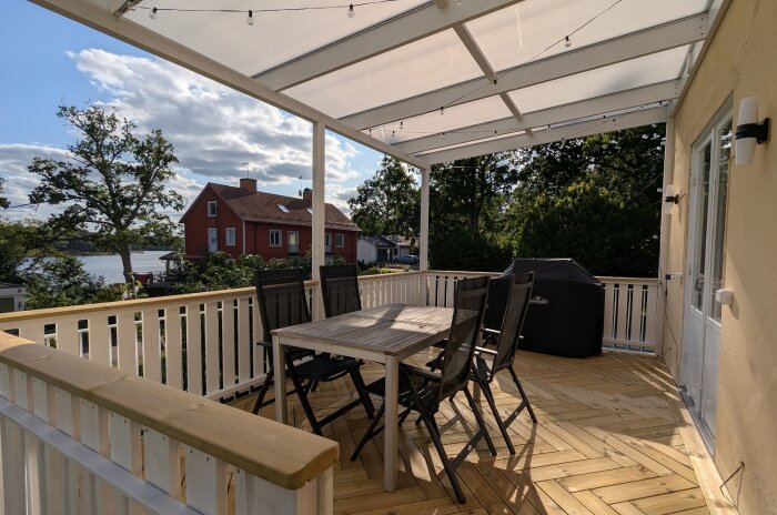 Altan byggd av trä med tak, utemöbler och en grill; omgiven av grönska och med utsikt mot ett rött hus och vattnet i bakgrunden.