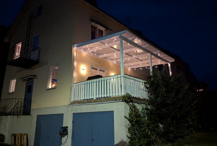 Nattvy av nybyggd altan med vit räckverk och belysning, placerad på ett tvåvåningshus.