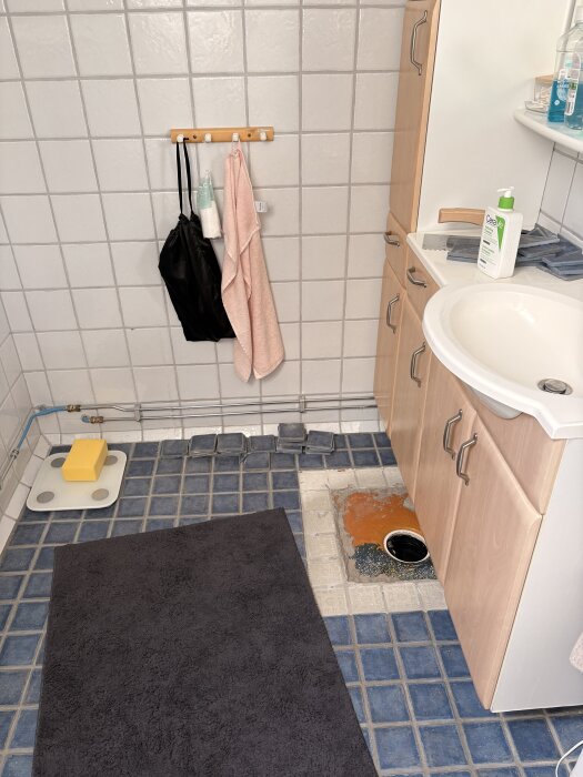 Badrum med blått kakelgolv, synlig golvbrunn vid sidan om ett vitt handfat, hagar och handdukshängare på väggen.