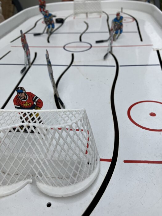 Bordshockeyspel med hockeyspelare i röda och blåa tröjor, en puck och målbur på ett vitt spelplan.