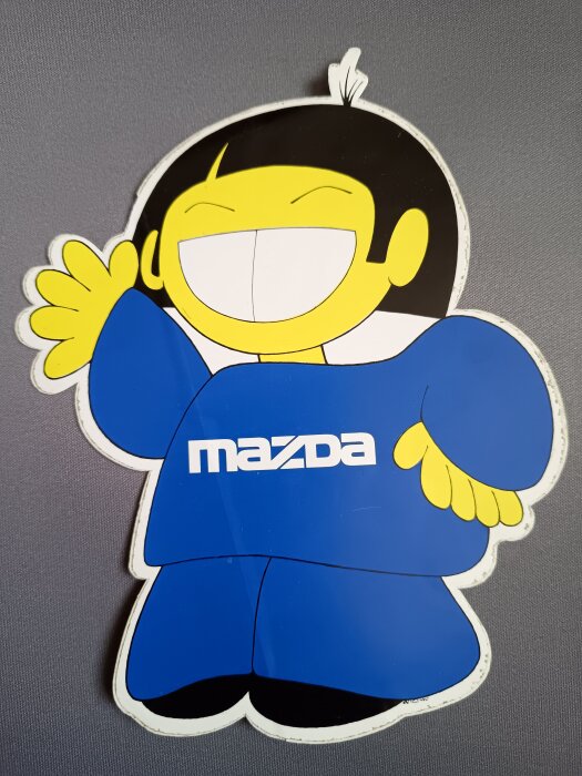 Tecknad figur med gul hud, leende ansikte och blå kläder med "Mazda" logga. Bild från Mazda-reklam från tidigt 80-tal.